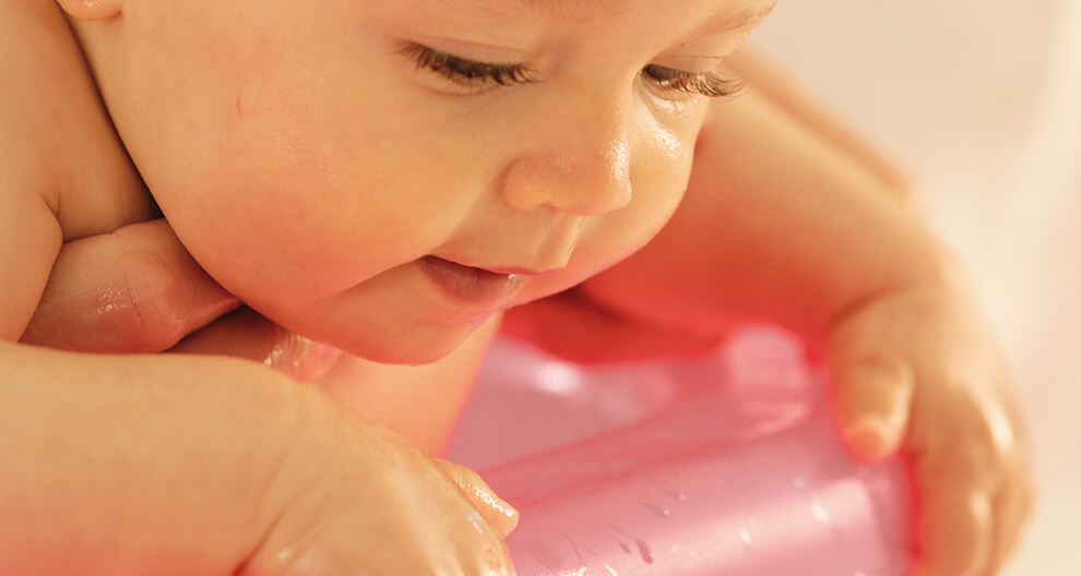 A close-up of a baby's face and hands, with water droplets on the skin, resting on a pink object.