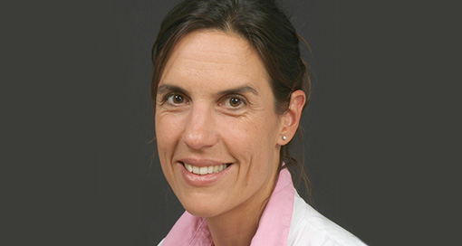 A woman with brown hair, wearing a pink shirt and white jacket, smiling at the camera.